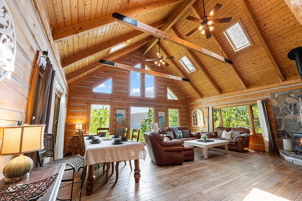 A cozy dining area with a large table set for a meal, comfortable leather sofas nearby, expansive windows framing mountain views, and a stone fireplace. The arrangement evokes warmth, togetherness, and scenic enjoyment.