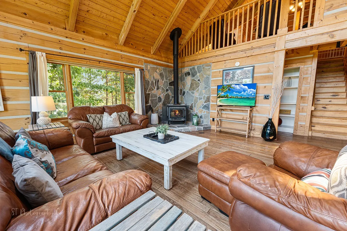 A bright, sunlit living area with comfortable leather sofas and a large wooden coffee table facing a beautiful stone fireplace. Generously sized windows let in natural light and showcase scenic outdoor vistas, while the arrangement encourages socializing and group activities.