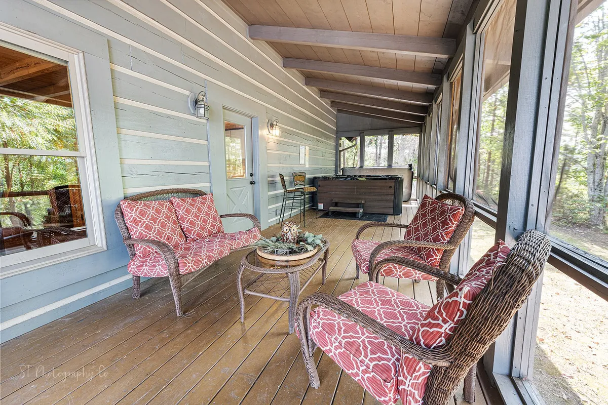 This exterior view highlights a screened-in porch featuring a hot tub, red patterned chairs, and a wooden table. Surrounded by forested scenery, the space exudes relaxation and social potential after a fun-filled day at local attractions.