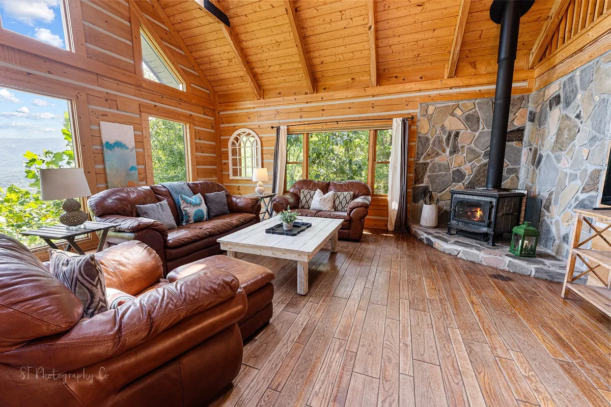 A welcoming open-concept living room with large leather sofas arranged around a stone wood-burning fireplace. Expansive windows draw in sunlight and frame picturesque mountain views. The space features warm wooden ceilings, and there's a sense of spaciousness ideal for gatherings or relaxing with family.