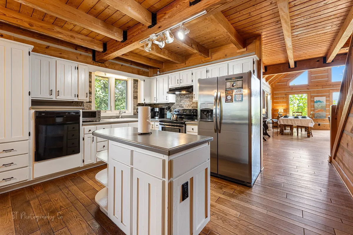 A bright, fully equipped kitchen featuring a central island, large stainless steel refrigerator, wooden ceiling beams, and a sunny window with views outside. Ample counter space and modern appliances are visible, showcasing an ideal environment for preparing meals for a group.